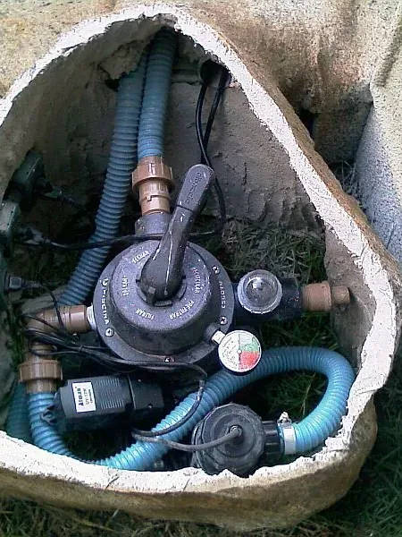 Pedra Western levantada no gramado, revelando registro de gás, mangueiras e caixa elétrica embaixo.